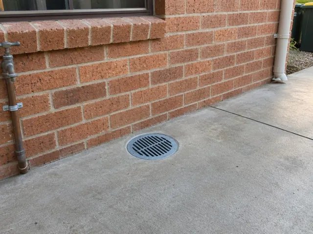 Round drain grate set in concrete beside a brick wall with a downpipe in Carramar